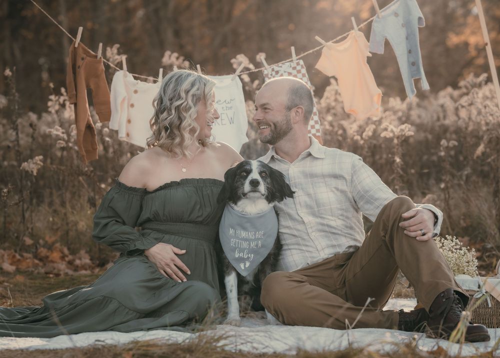 A couple sits on a blanket outdoors with their dog, surrounded by autumn foliage and clotheslines with laundry.