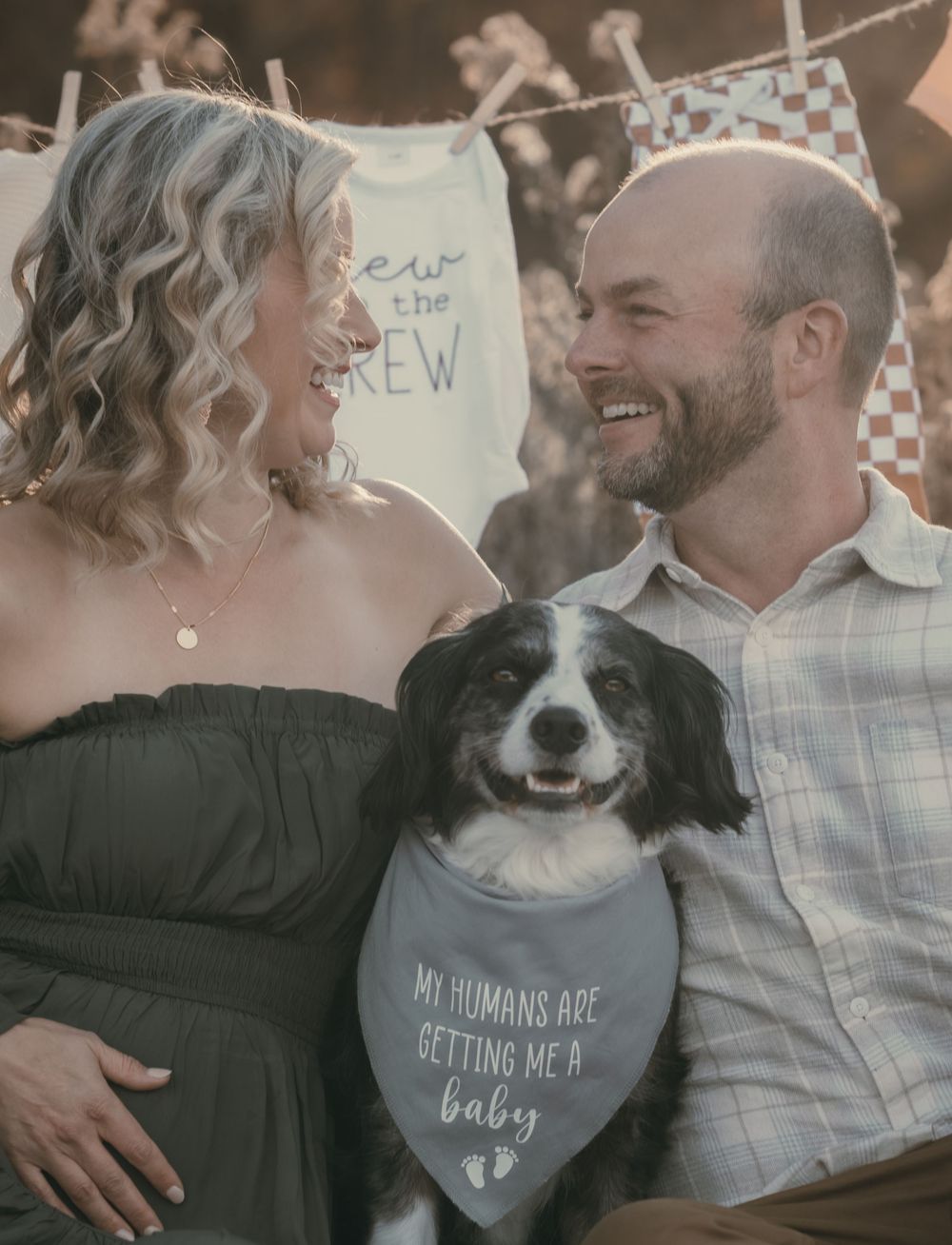 A couple poses with their smiling Border Collie wearing a bandana announcing a new baby on the way.