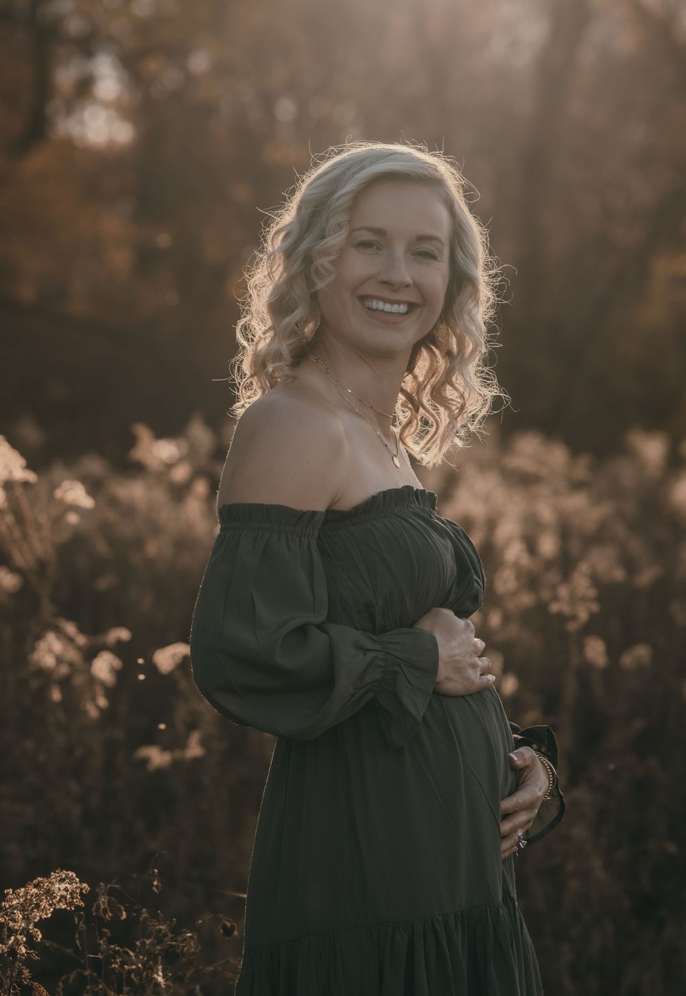 A woman in an olive green off-shoulder dress poses in a sunlit field during golden hour.