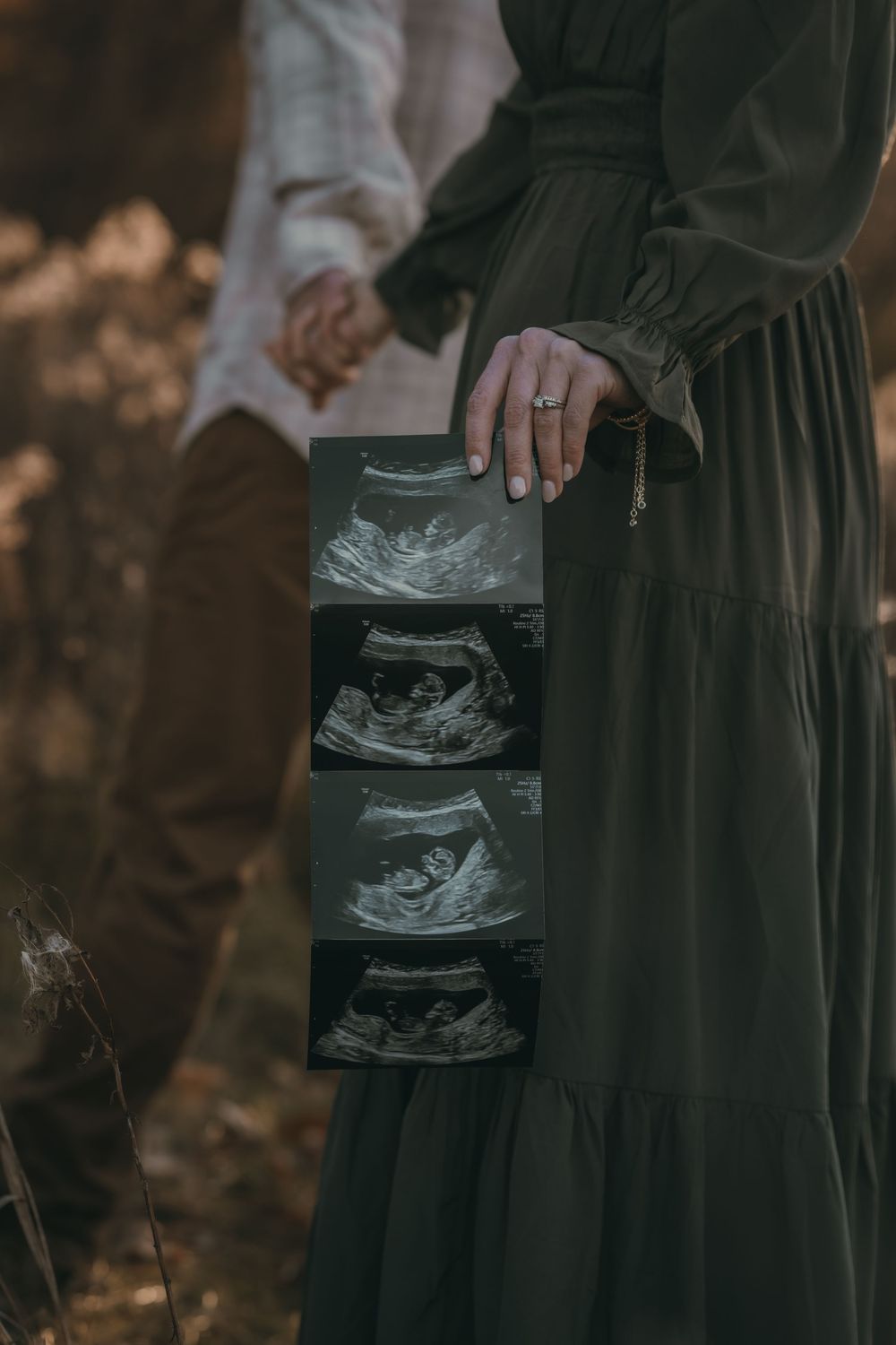 Someone in a dark flowing dress holds ultrasound images while holding hands with another person in a romantic outdoor setting.