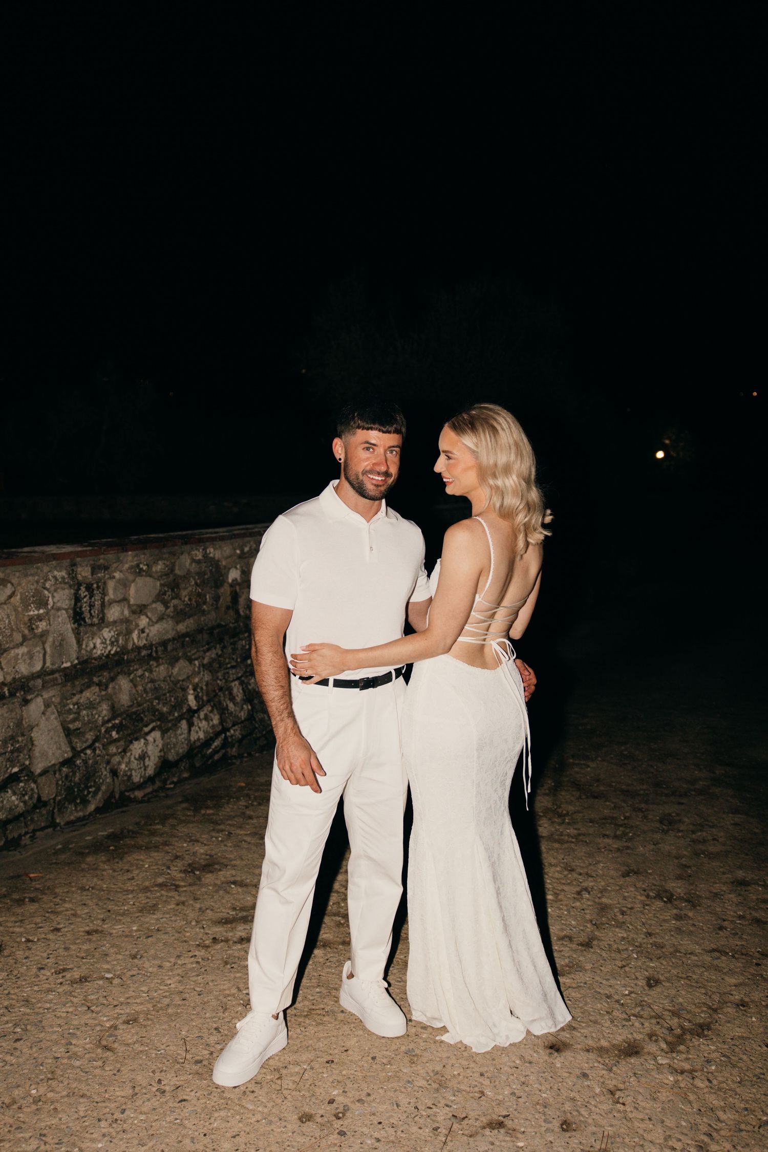 Elegant couple in all white attire posing at night against stone wall.