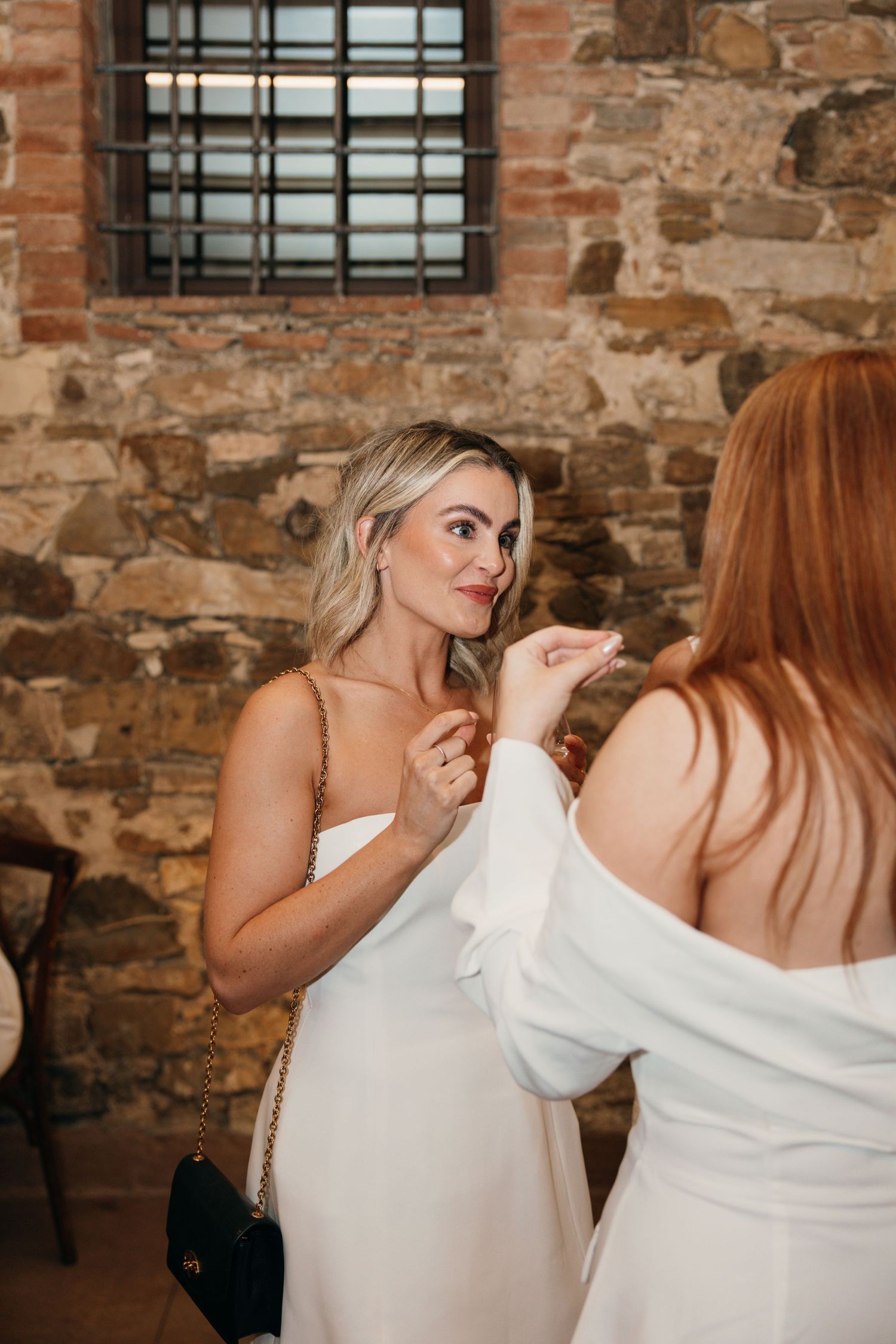 Wedding guests in white dresses having an animated conversation against a rustic stone wall backdrop.