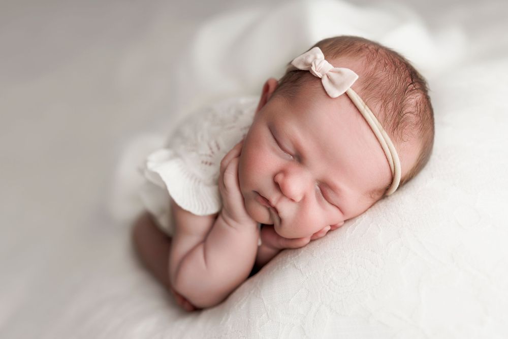 A sleeping newborn baby wearing a delicate white bow headband rests peacefully on soft white fabric.
