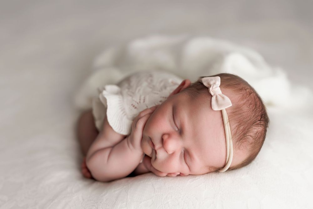 Sleeping newborn baby wearing delicate white headband lies peacefully on soft textured fabric.