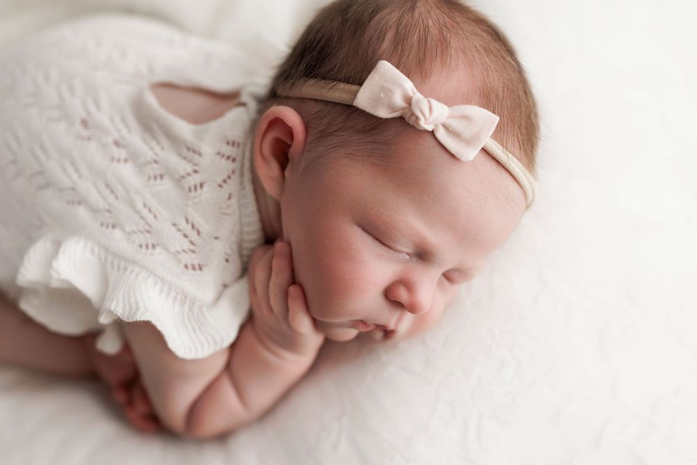 Sweet sleeping newborn in white lace dress and bow headband rests peacefully against soft white backdrop.