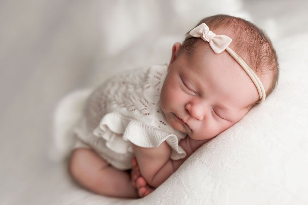 Sleeping newborn baby wearing white ruffled outfit and bow headband while resting on soft white blanket.