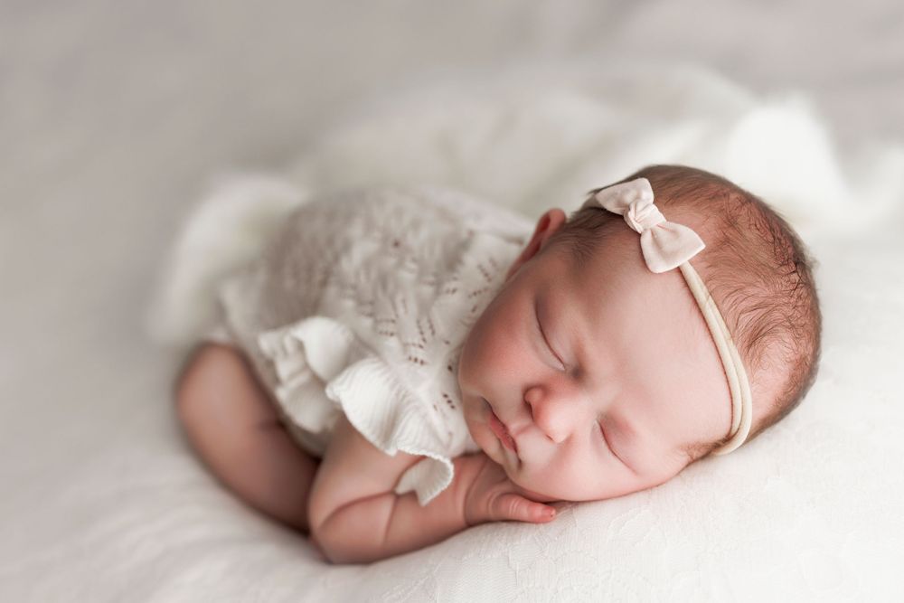 A peaceful sleeping newborn wearing a white lace dress and delicate bow headband rests on soft fabric.