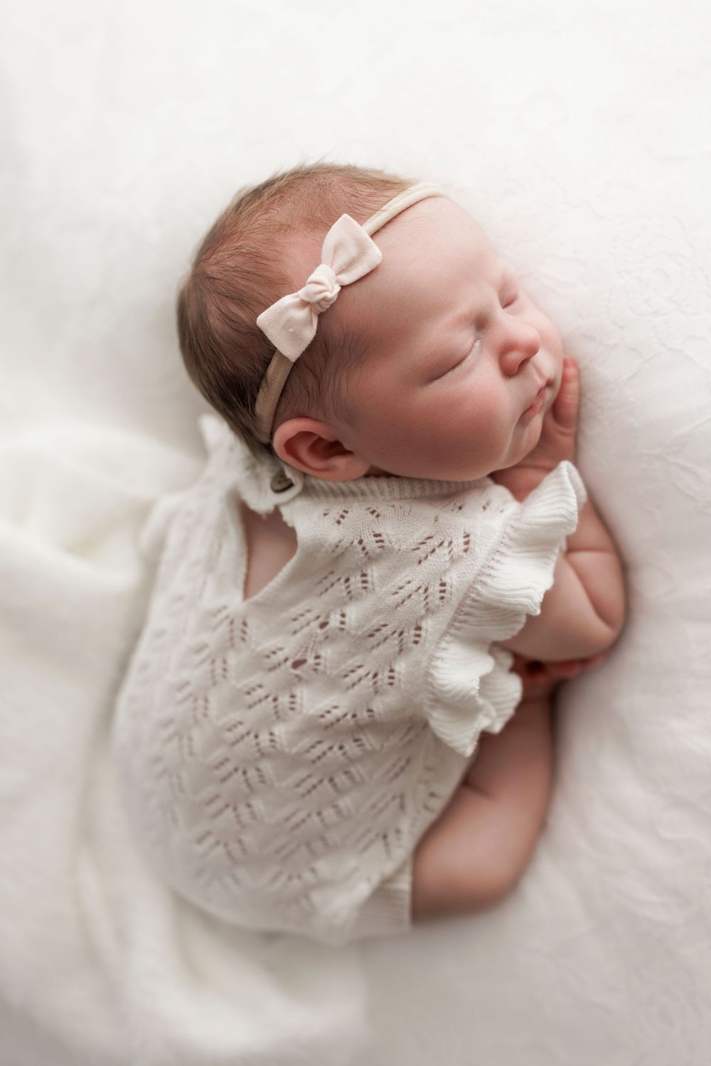 Sleeping baby wearing white knit dress and bow headband lies peacefully on soft white blanket.