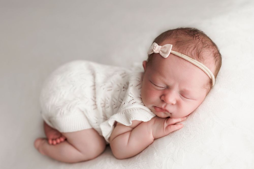 Newborn baby sleeping peacefully in white wrap wearing delicate bow headband on light backdrop.