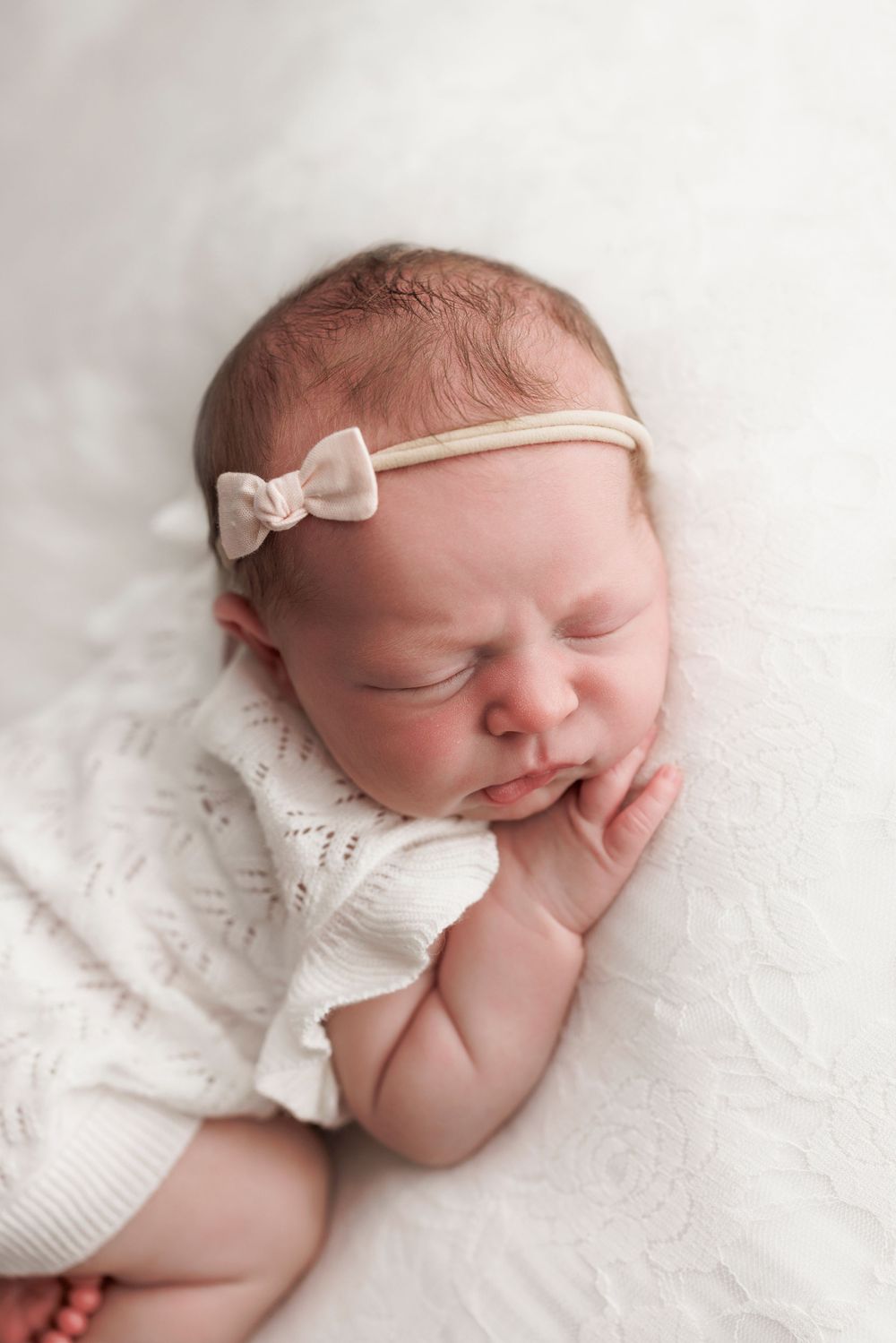 Sleeping newborn baby wearing a white knit outfit and delicate bow headband rests peacefully on soft white fabric.