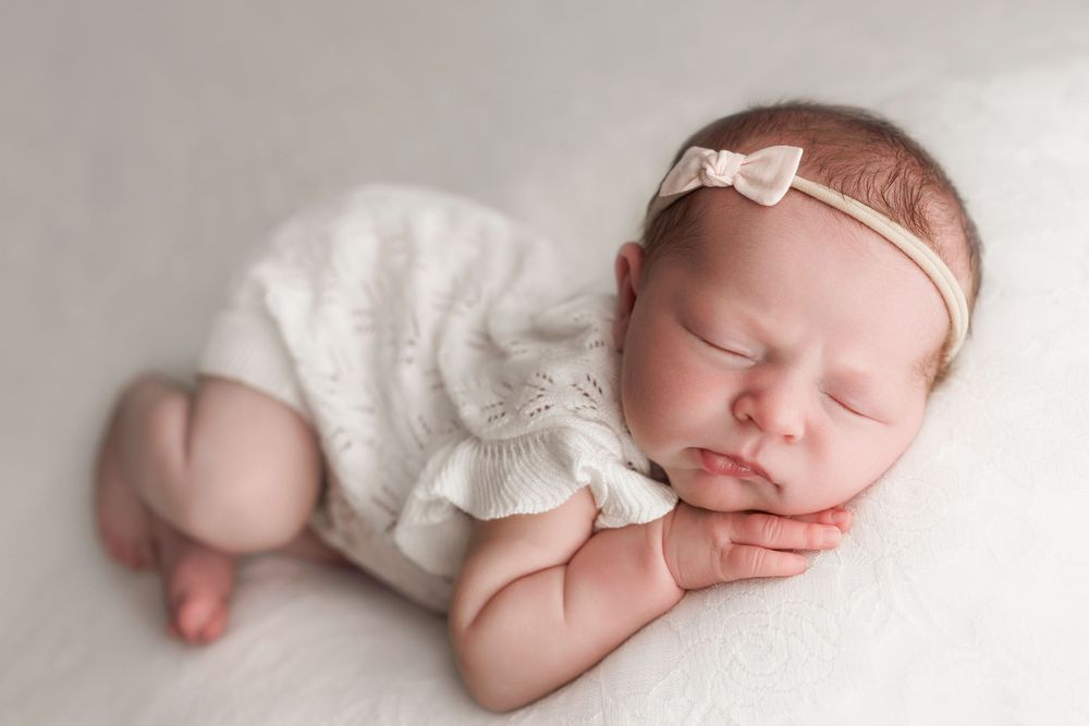 Sleeping newborn in white knit outfit lying on side with small bow headband on light background.