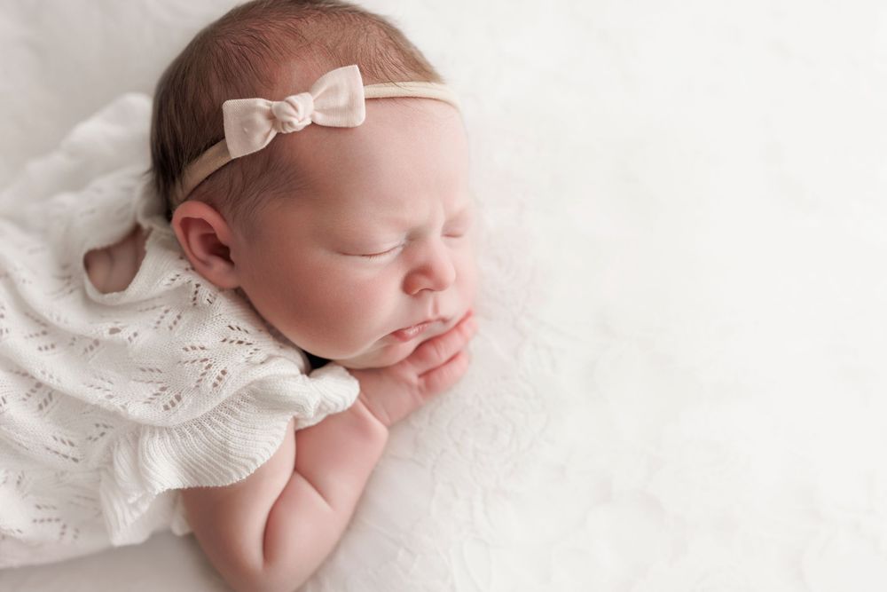 Peaceful sleeping infant wearing lace dress and bow headband on white backdrop.