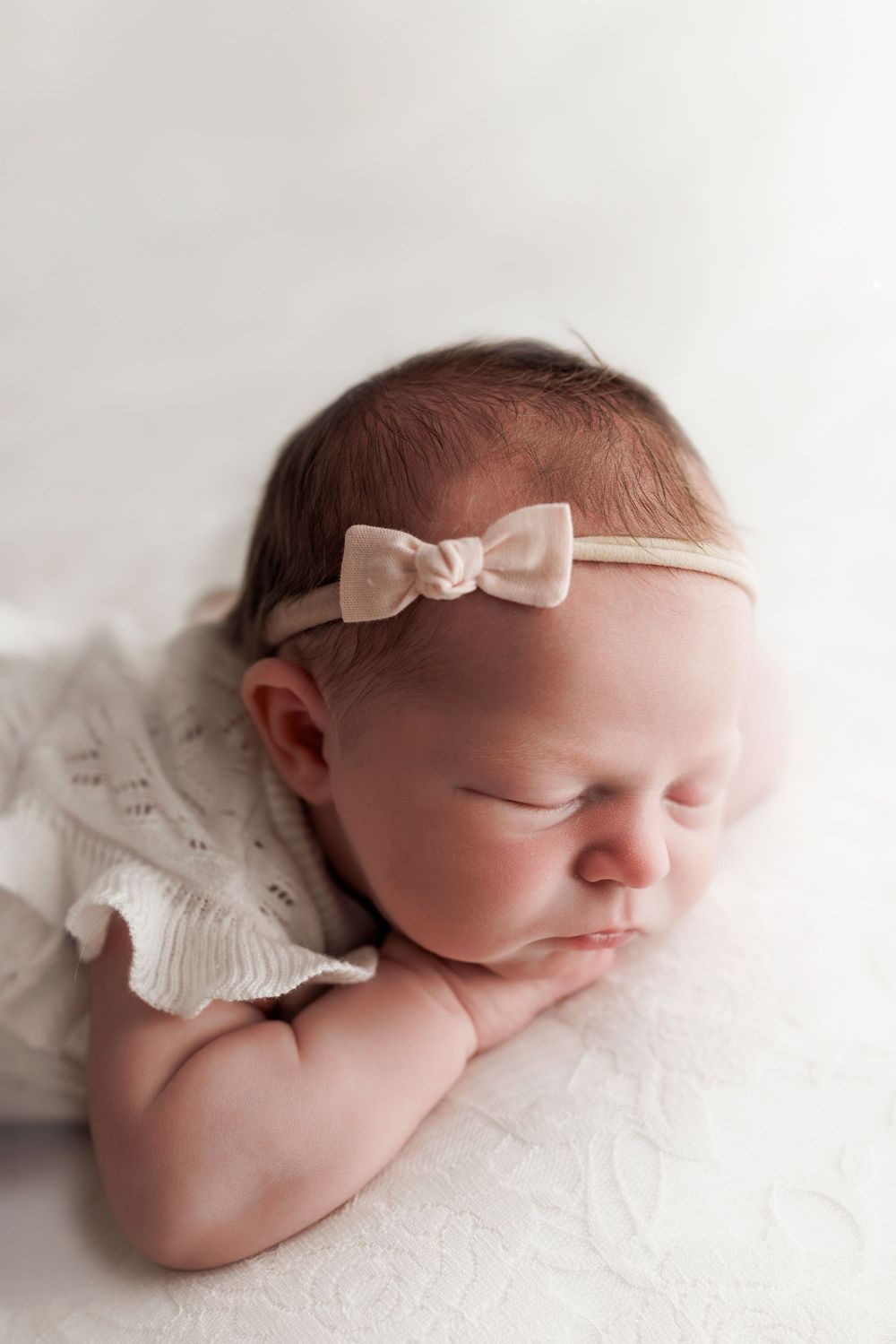 Peaceful sleeping newborn wearing white lace outfit and delicate bow headband on soft white backdrop.