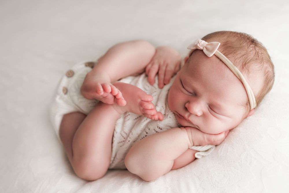 Resting newborn in white outfit with delicate headband against soft white background.