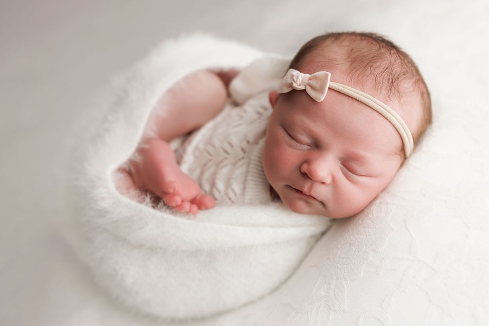 A sleeping newborn baby swaddled in soft white fabric with a delicate white headband rests peacefully.
