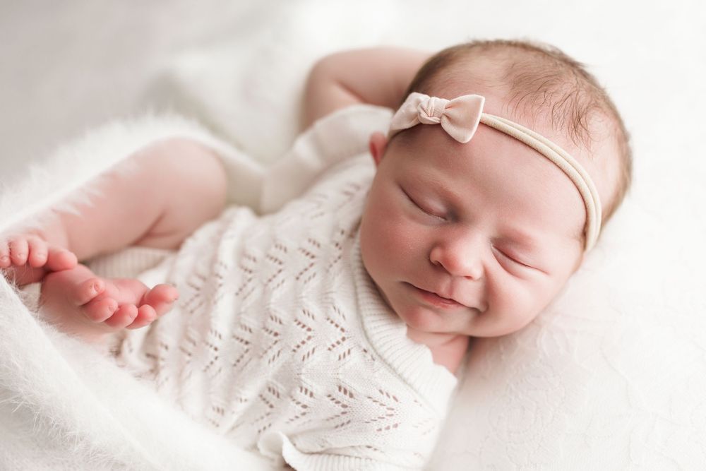 Sleeping newborn baby wearing a white knit sweater and delicate bow headband rests peacefully on soft blanket.