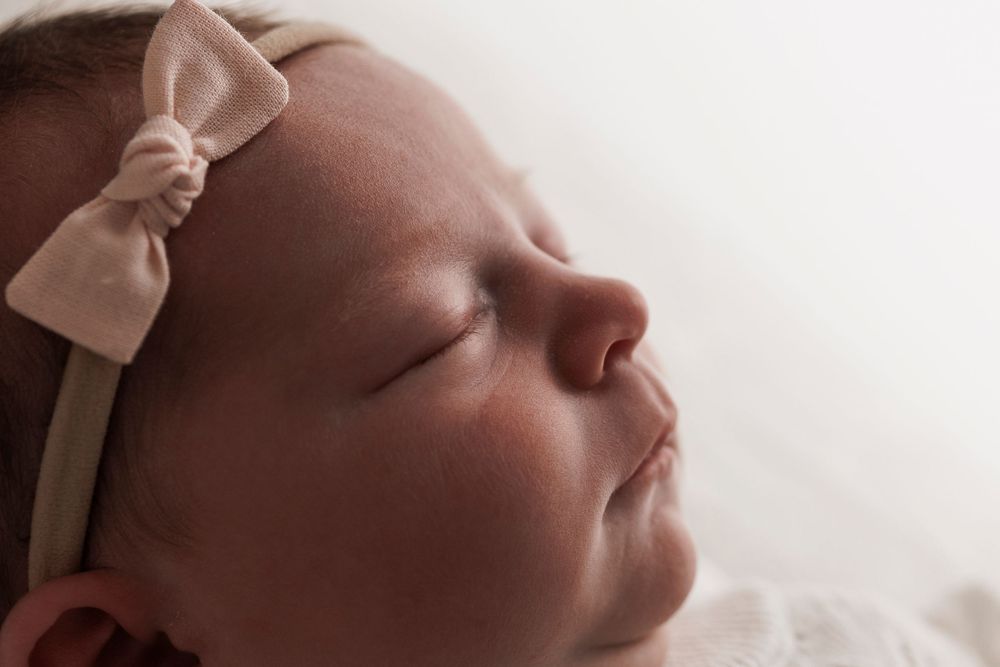 A sleeping newborn wearing a delicate pink bow headband rests peacefully in a close-up portrait.