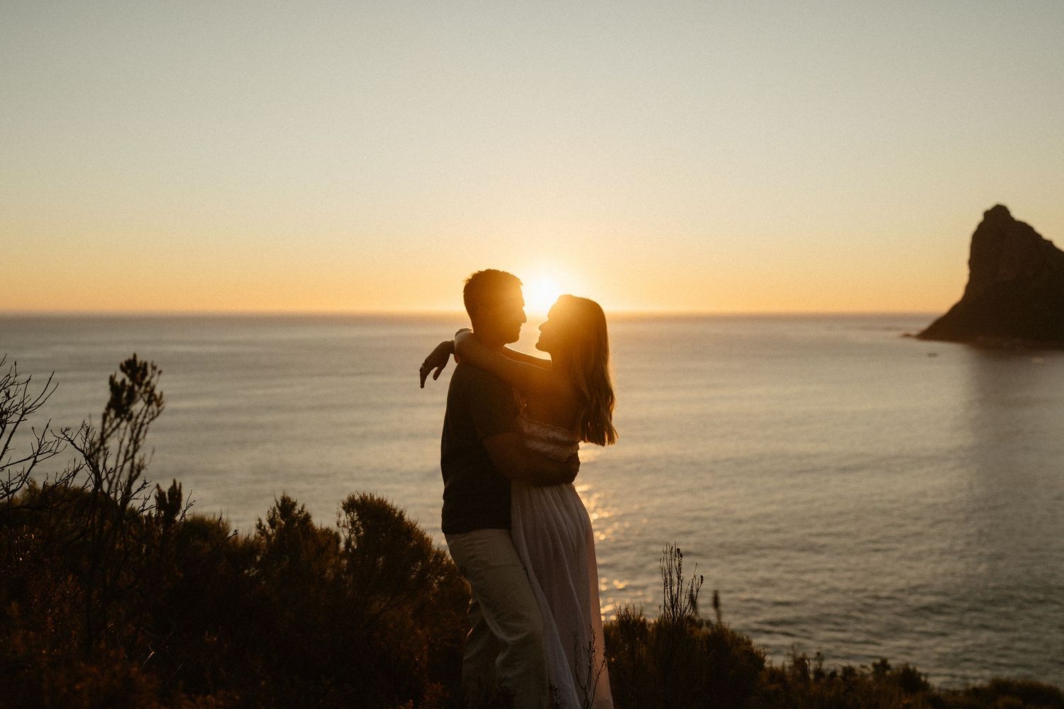 A couple in dark clothing embraces while standing on rocky coast with mountain peak in background at sunset.