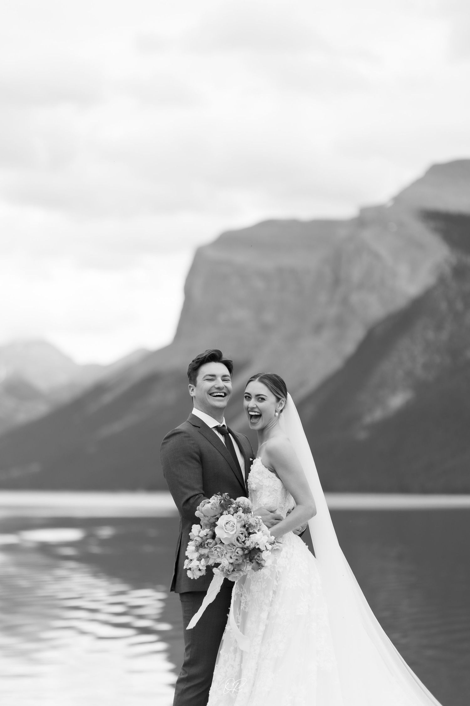 A newlywed couple sharing a joyful moment by a scenic mountain lake with dramatic peaks in the background.
