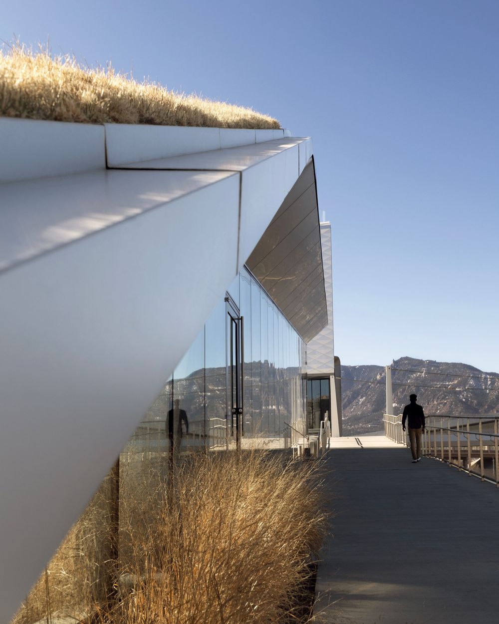Olympic and Paralympic Museum modern architectural walkway with glass walls and angular white roof against mountain backdrop and blue sky.