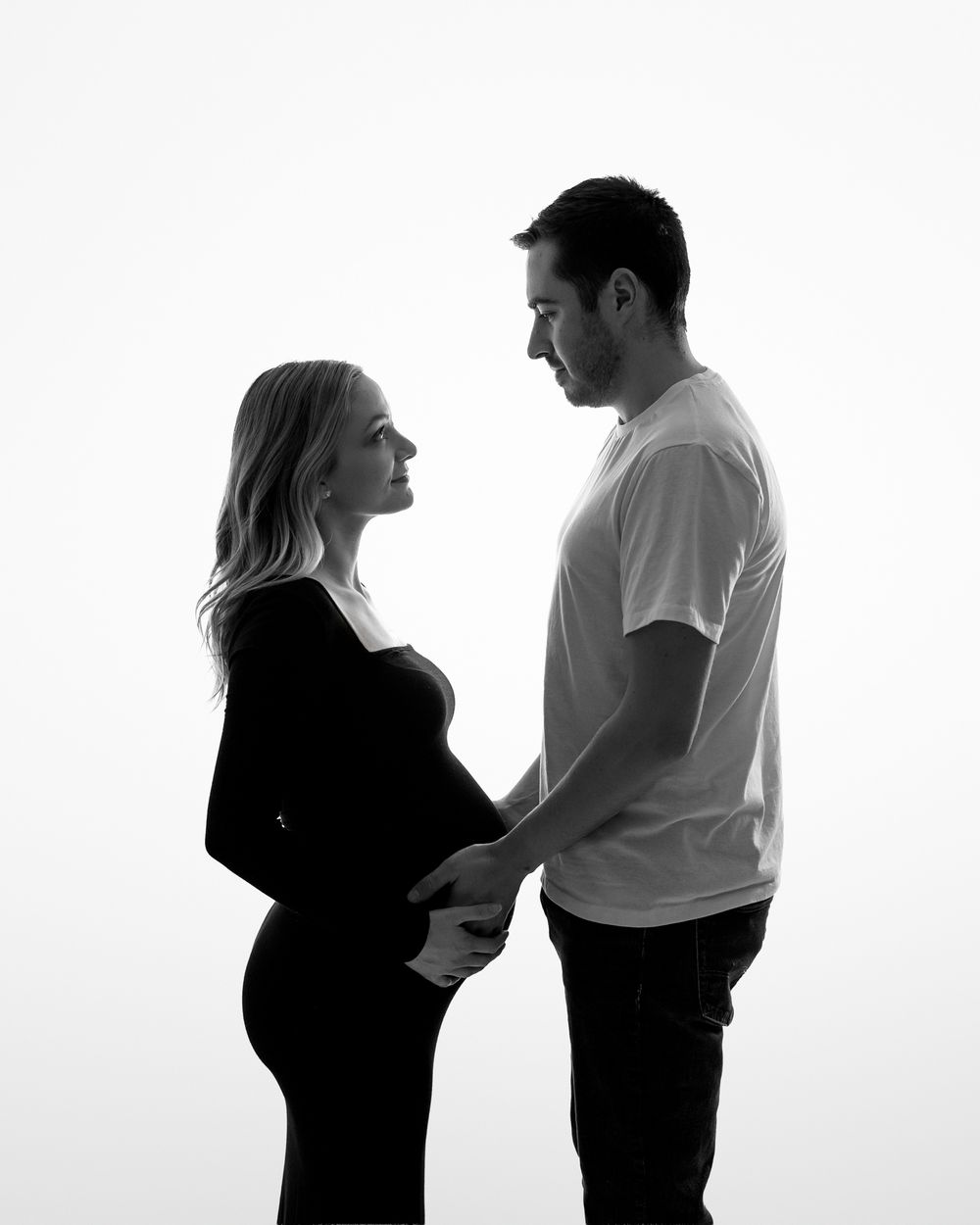 Black and white silhouette portrait of an expectant couple sharing an intimate moment against a bright background.
