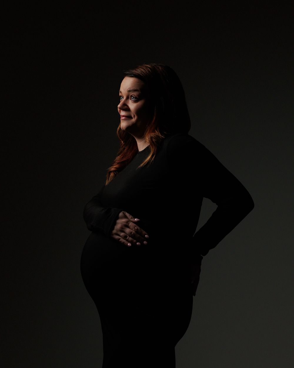 Dramatic silhouette maternity portrait showing an expectant mother in a black dress against a dark background.