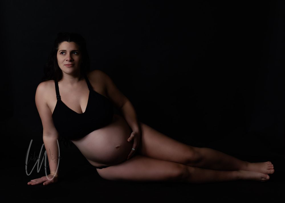 A person in black swimwear poses gracefully in a dramatic low-key maternity photo against a dark background.