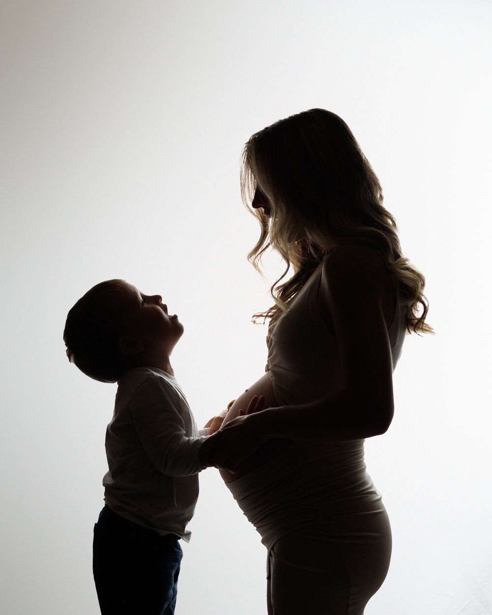 Silhouetted side view of young child touching pregnant belly against white background.