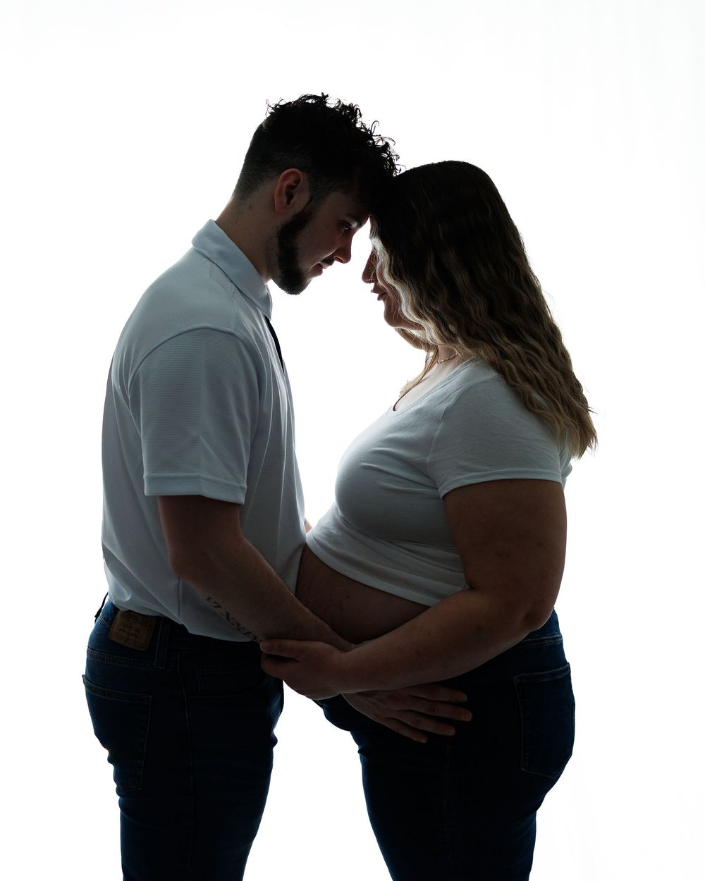 Silhouetted couple shares an intimate moment during maternity photoshoot against white background.