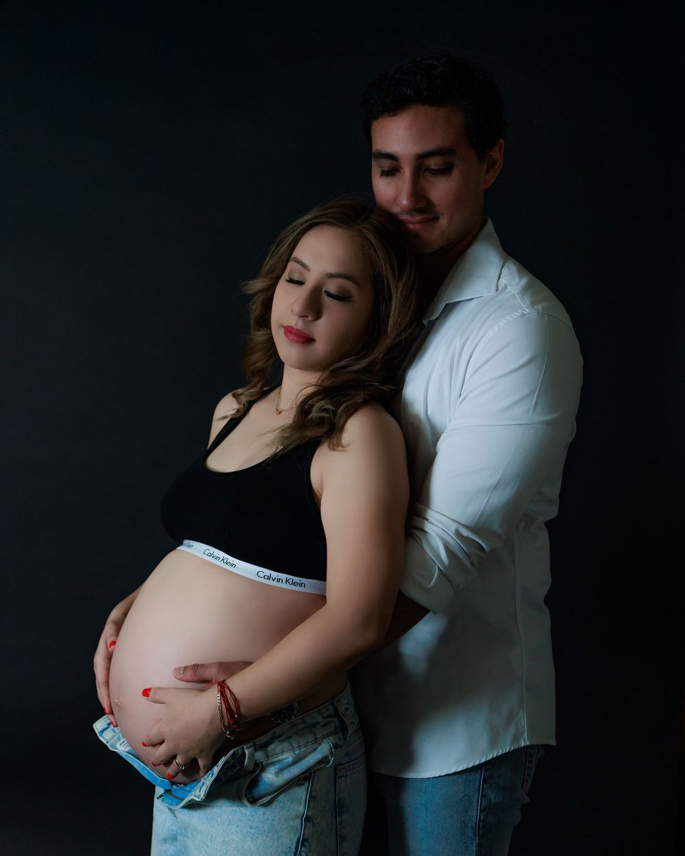 A romantic maternity photo shoot shows an intimate embrace against a dark background.