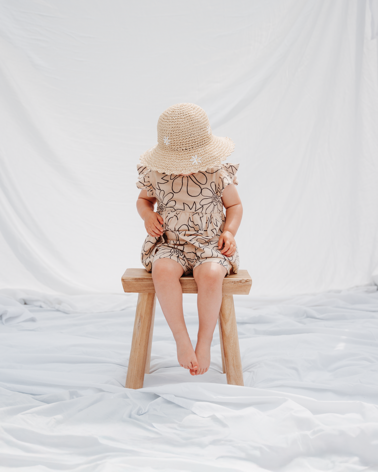 Small child sitting on wooden stool wearing patterned outfit and straw hat against white backdrop.