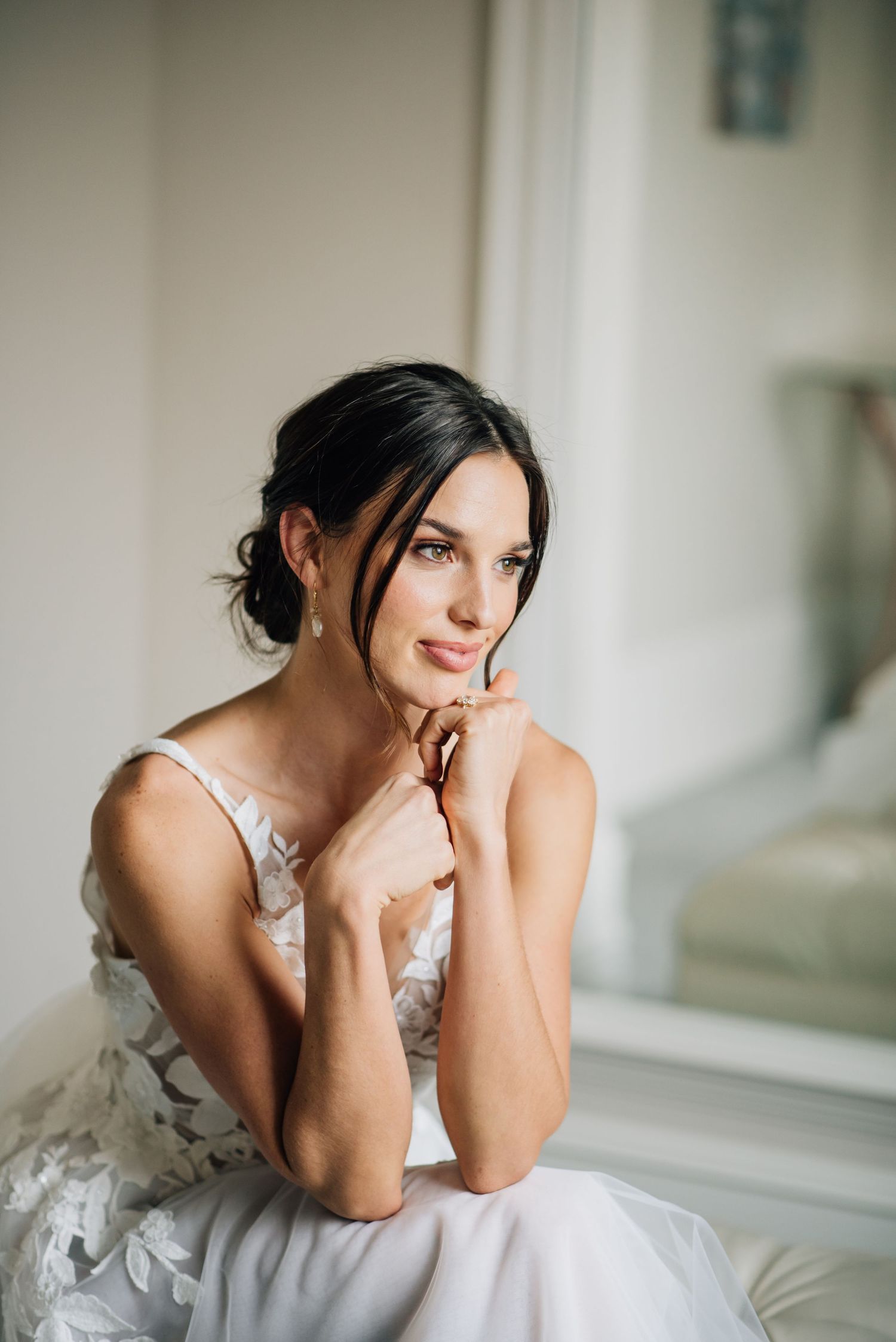 Elegant bride in flowing white gown sits with coral and pink peony bouquet in bright white reception room with gold chairs.