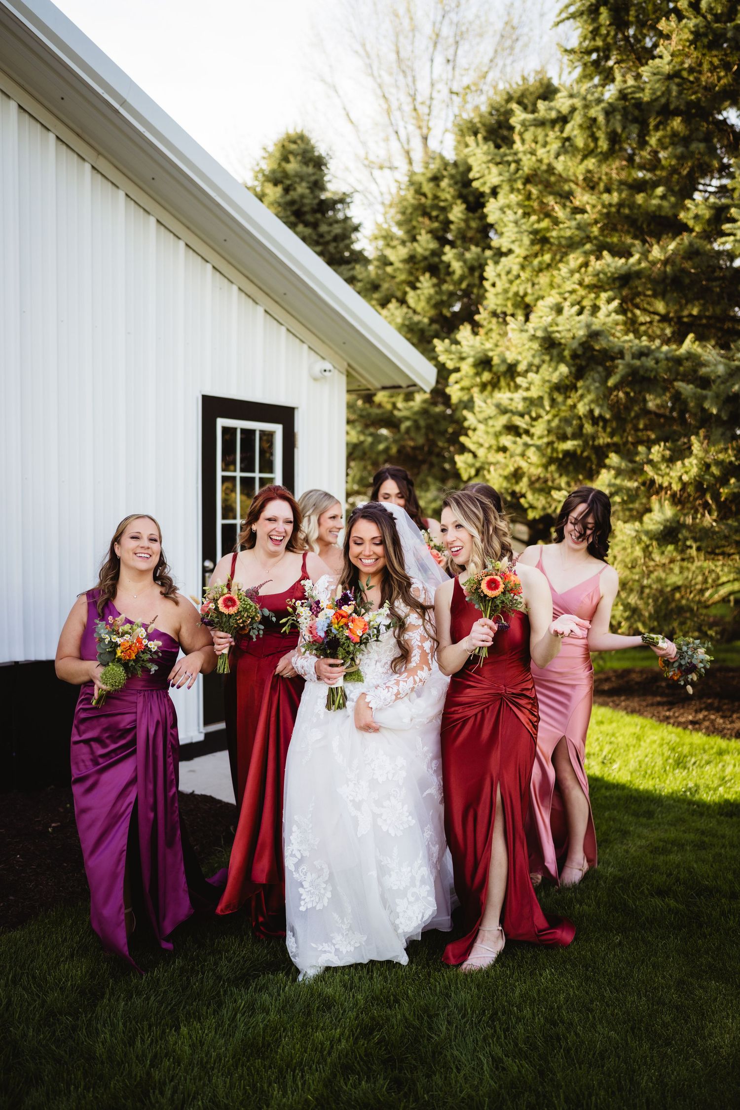Wedding party in shades of burgundy and pink dresses pose and laugh together on lawn outside white building.
