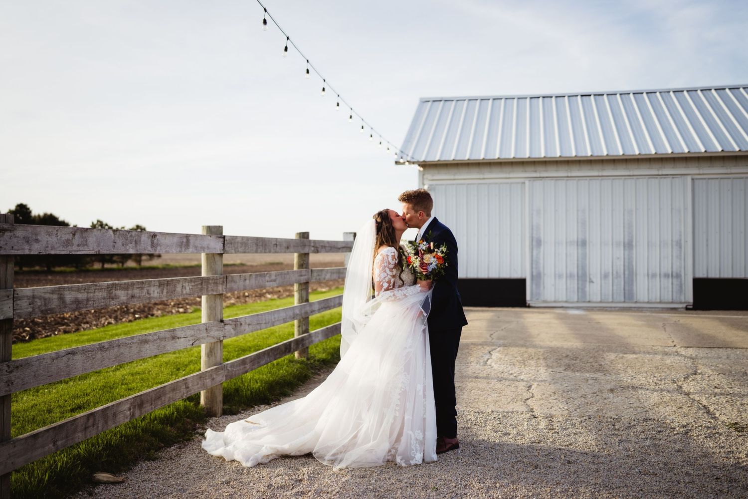 A bride and groom share a romantic moment by a rustic white barn and wooden fence on their wedding day.
