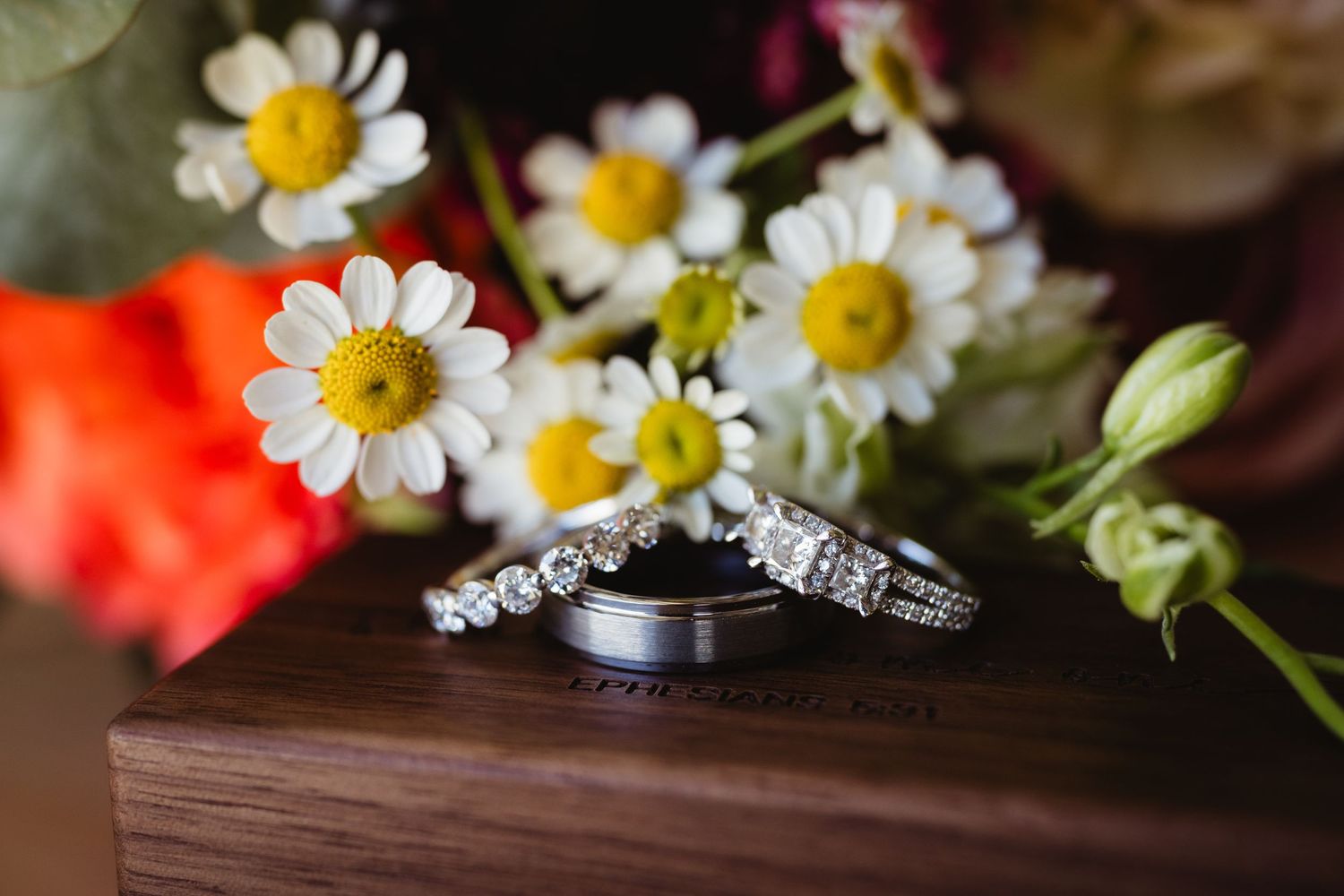 Wedding rings displayed on a wooden surface with white daisies and red flowers in the background.