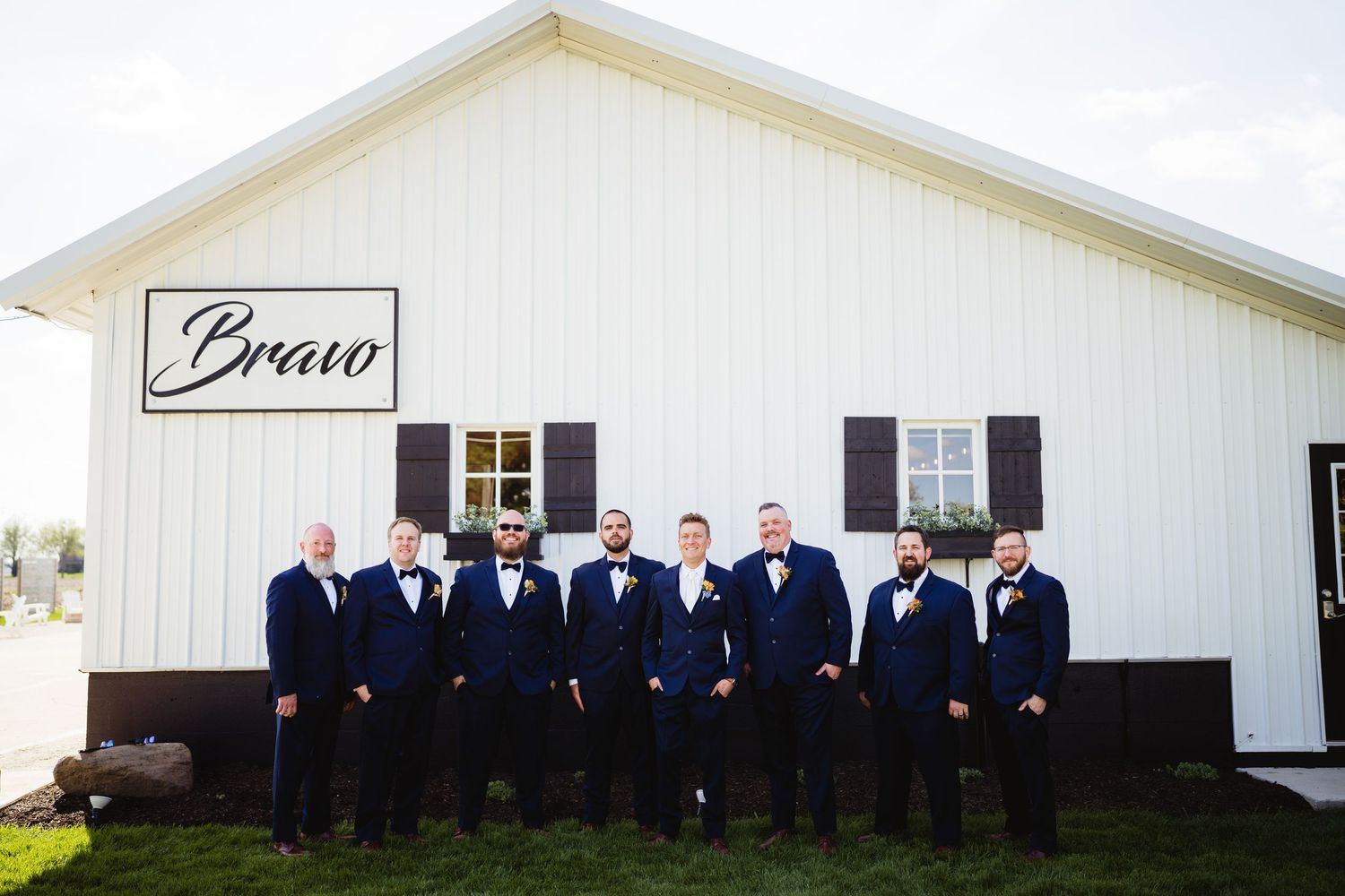 A group of groomsmen in navy suits and bow ties stand in front of a white wedding venue with a Bravo sign.