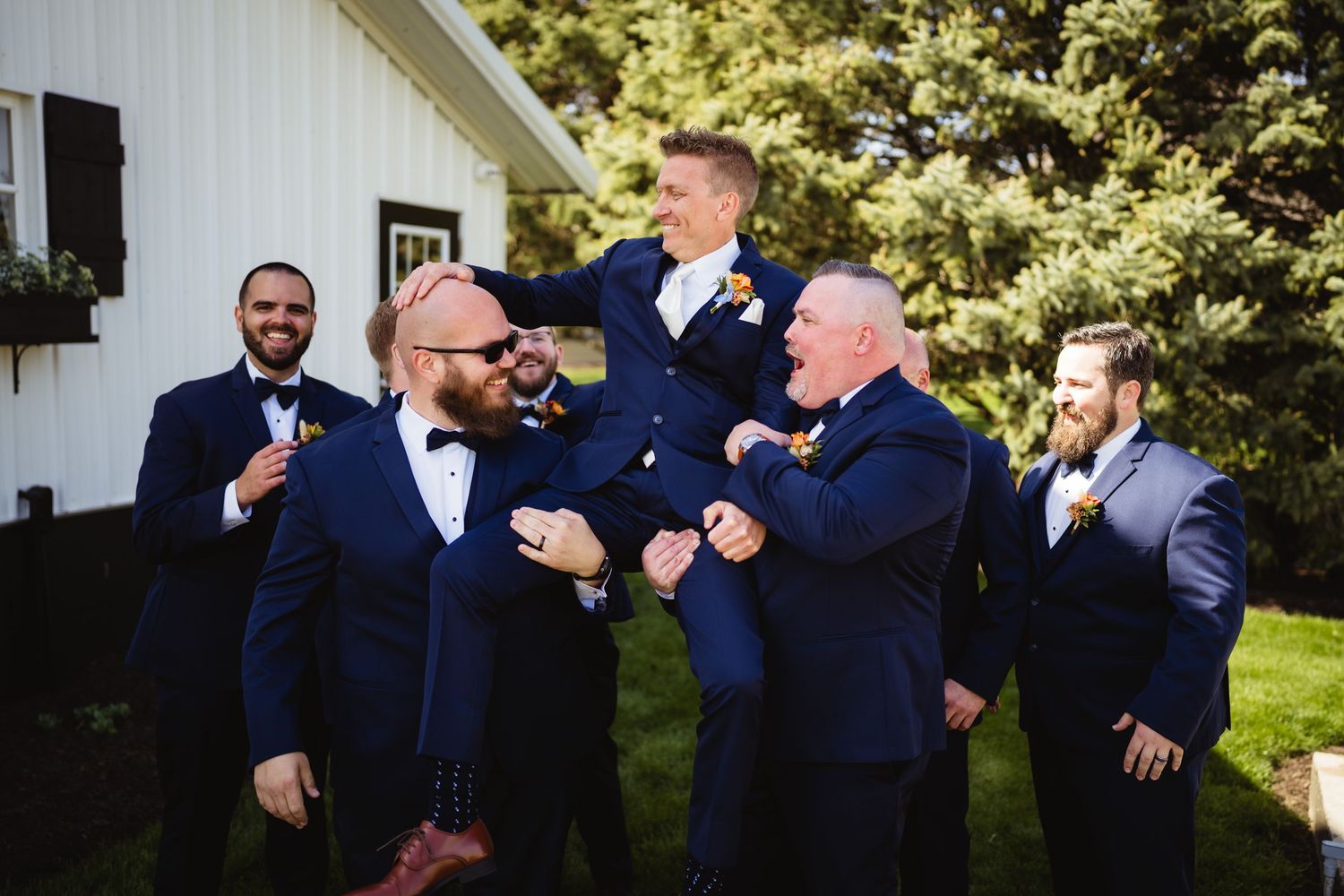 A group of men in navy blue suits laugh and celebrate together at an outdoor wedding event.