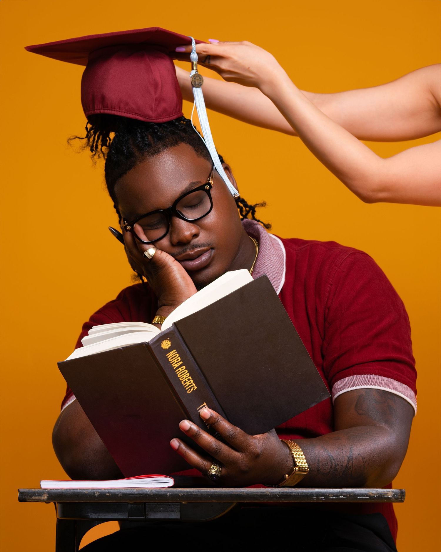 A student in a burgundy graduation cap and sweater reads a book while someone adjusts their cap against an orange background.