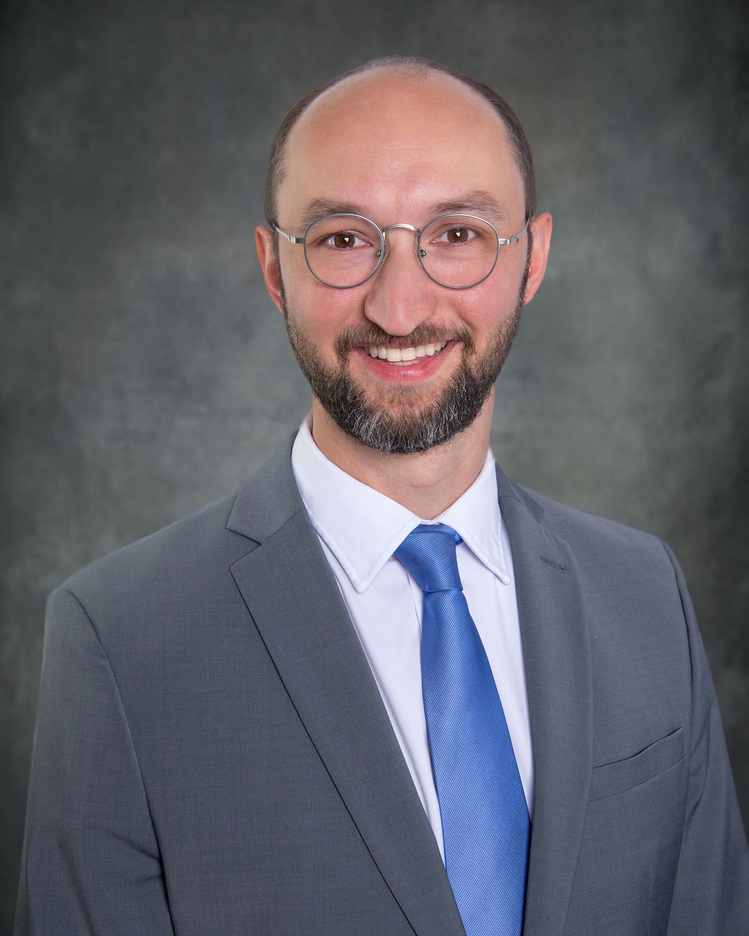 Professional headshot portrait of someone in a gray suit and blue tie with glasses and a neatly trimmed beard.