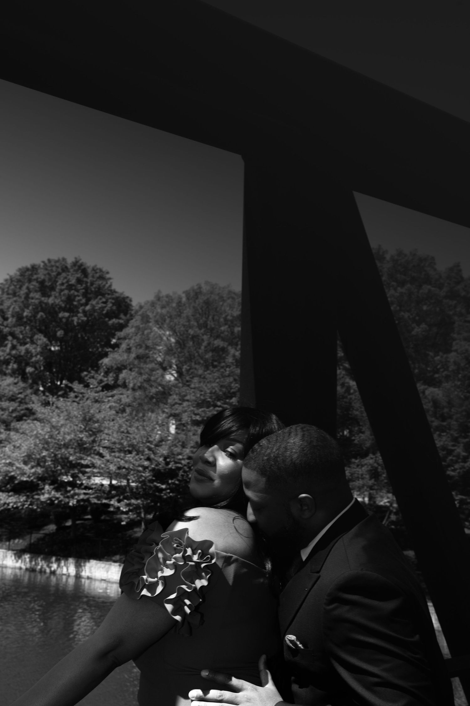 A romantic black and white photograph of a couple in formal attire sharing an intimate moment on a bridge over water.