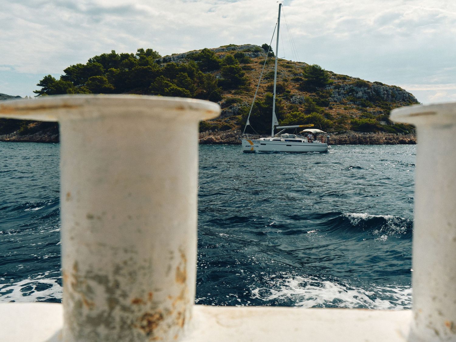 View of a sailboat anchored near a rocky island seen through white boat railings on choppy blue water.
