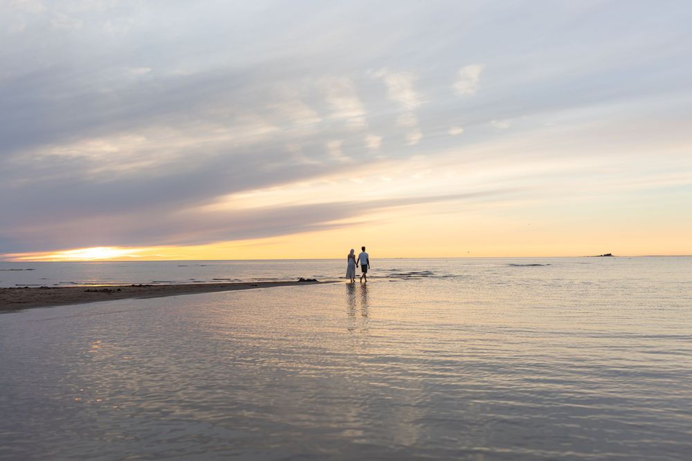 Two silhouettes stand together on a beach sandbar during sunset over calm ocean waters.