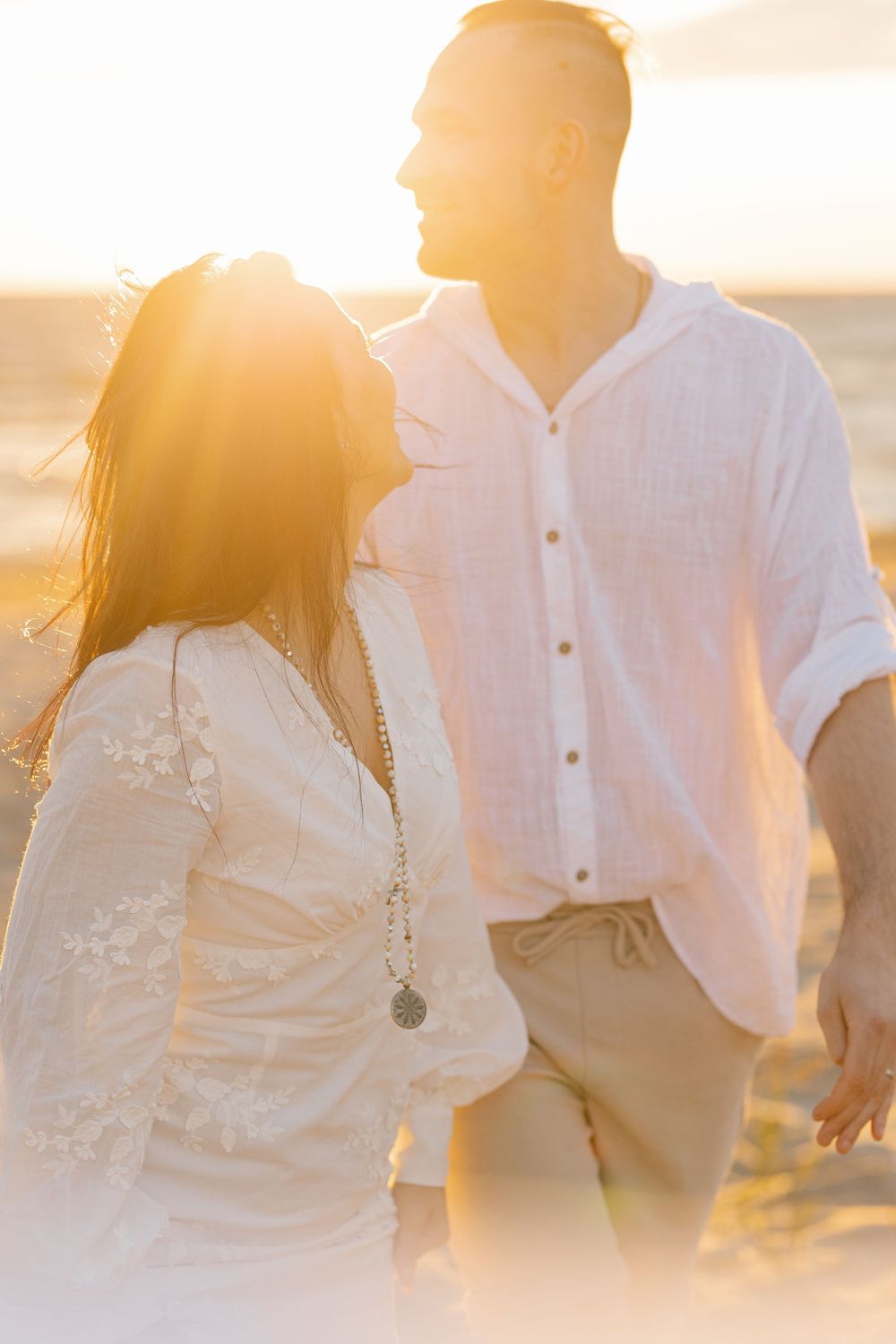 A couple in white attire embraces during golden hour on the beach with sunlight creating a dreamy atmosphere.