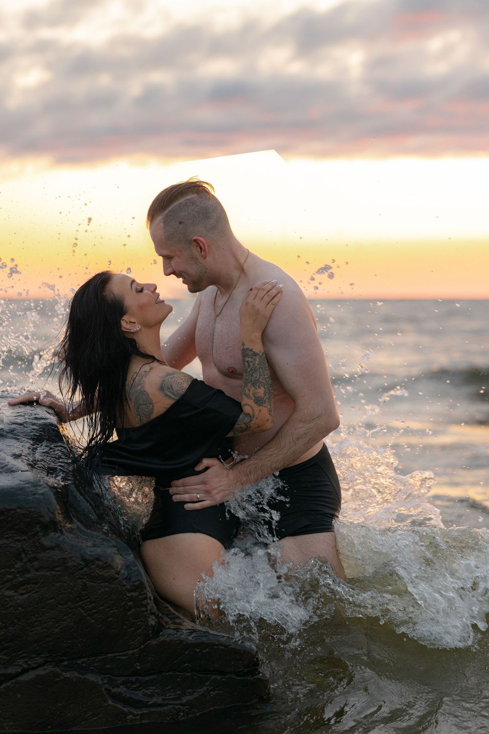 Couple playing in ocean waves during sunset photoshoot wearing black swimwear.