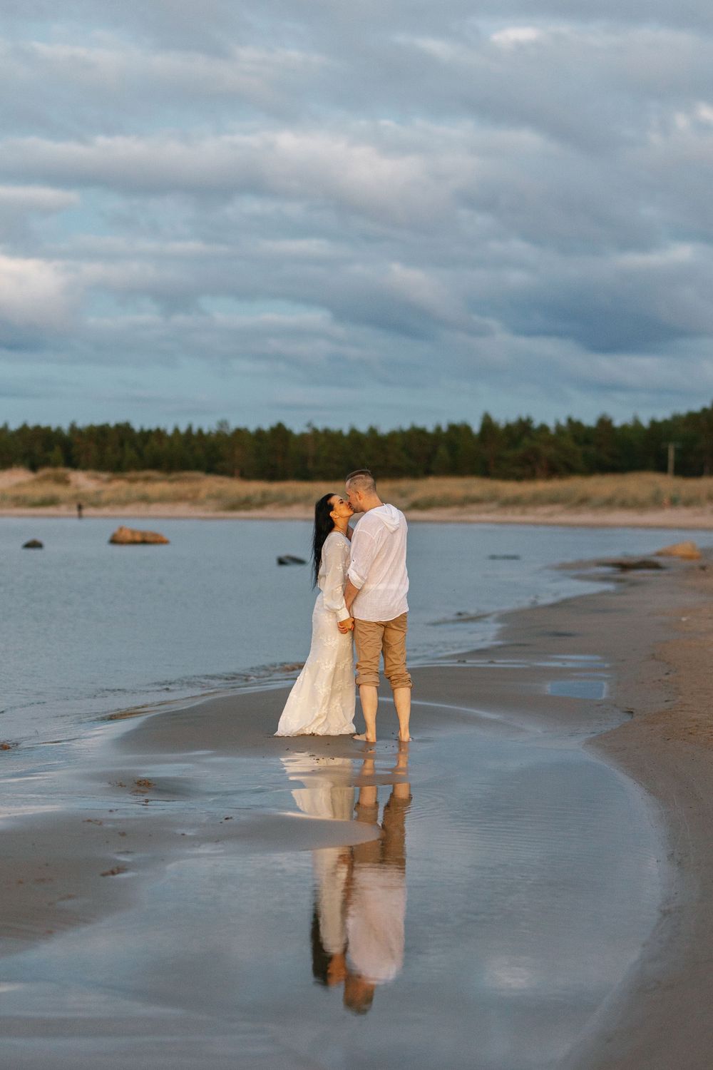 A couple in wedding attire embrace on a foggy beach at sunset with their reflection visible in the wet sand.