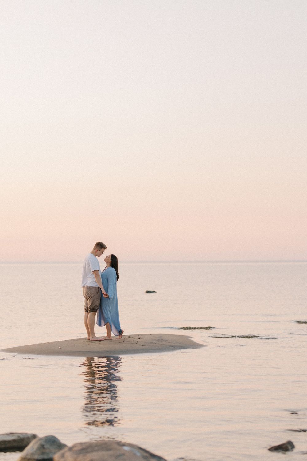 A couple stands together on a sandy beach during sunset with calm waters and rocks in the background.