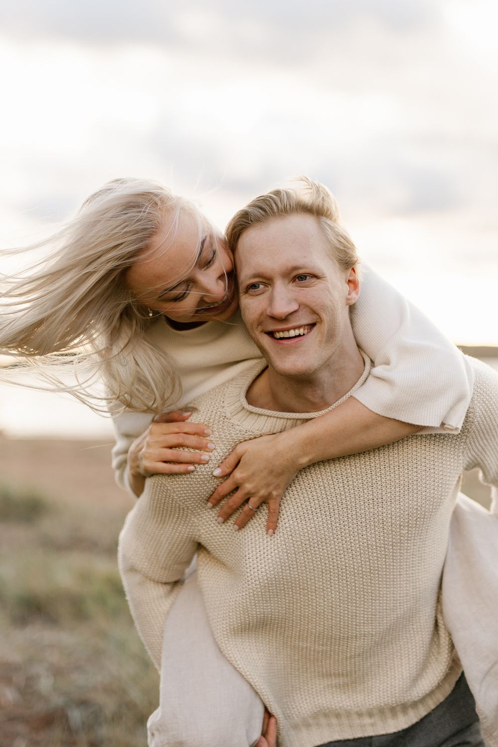 A couple shares a joyful piggyback moment outdoors wearing cozy cream-colored sweaters at sunset.
