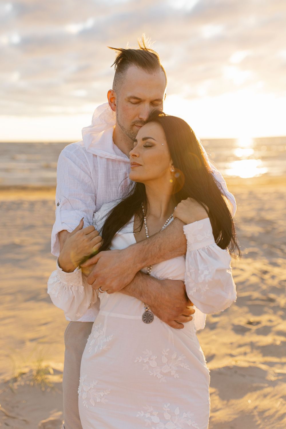 A couple embraces on a sandy beach at sunset wearing white outfits against the golden sky.