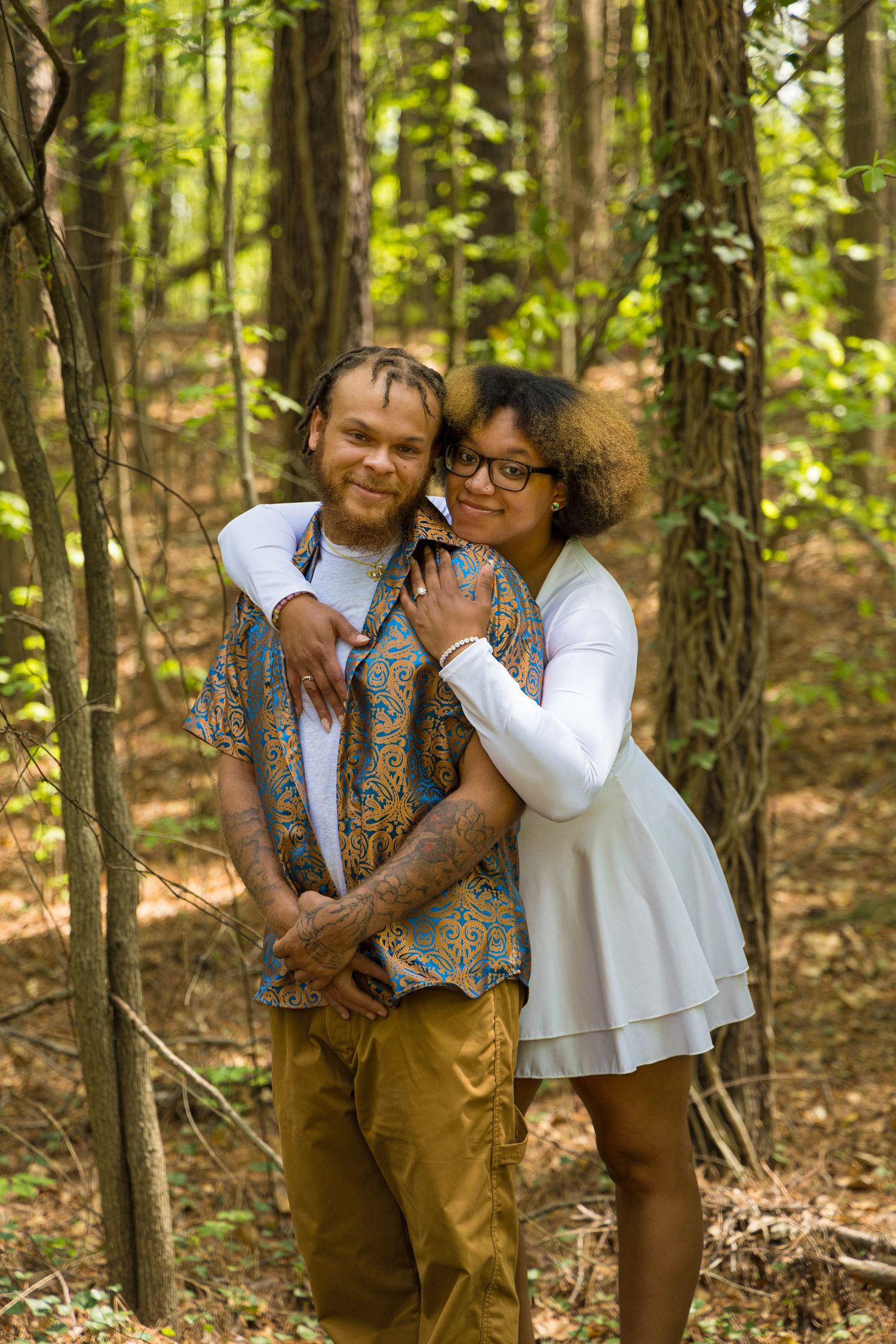A couple embraces in a sunlit forest setting wearing coordinated outfits with his patterned vest and her white dress.
