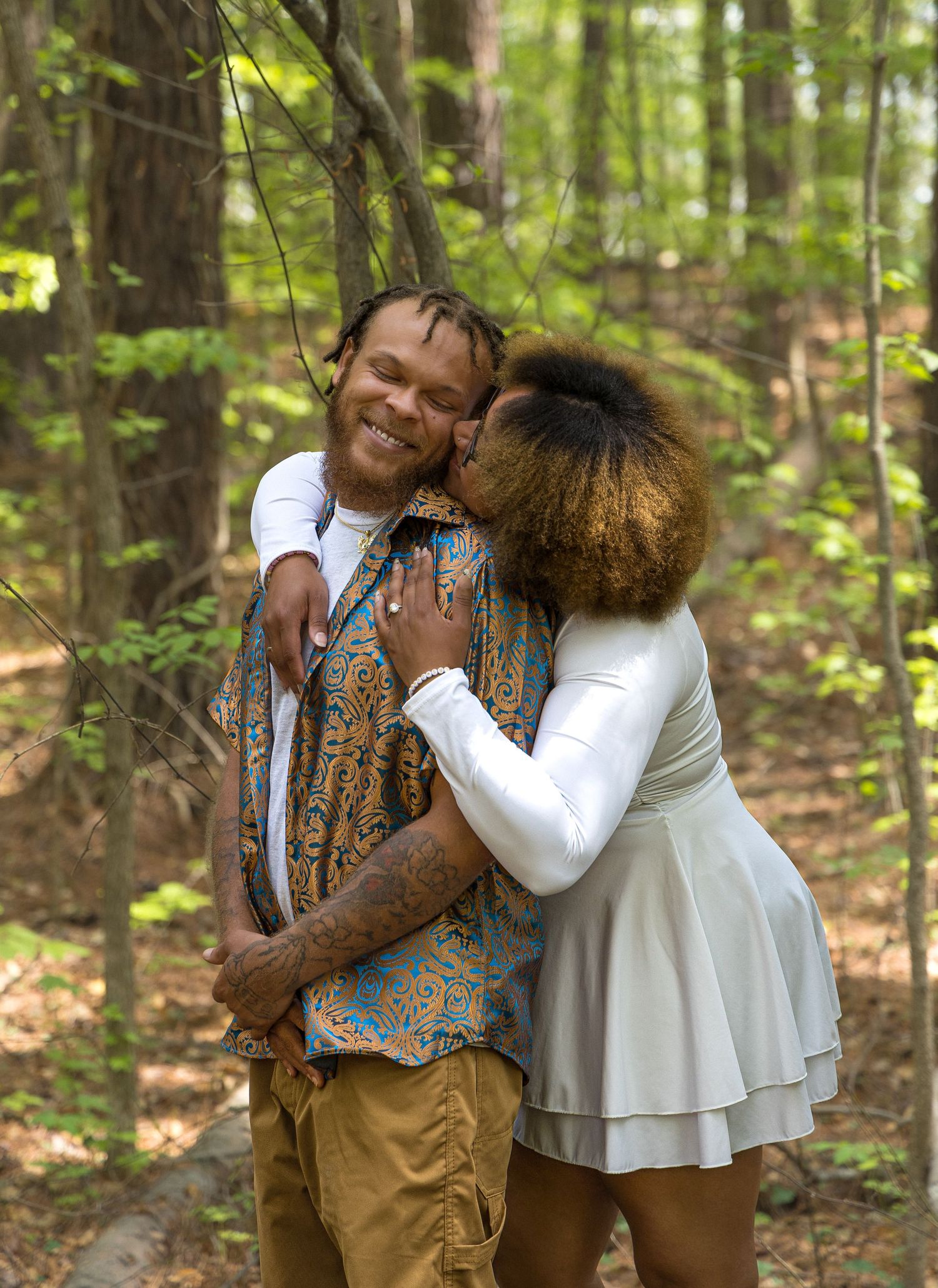 Two people in casual attire share an intimate embrace while smiling in a sunlit wooded forest setting.